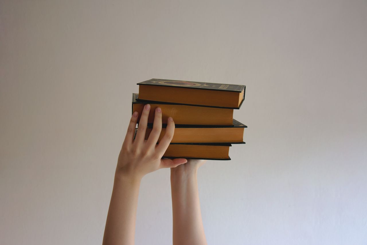 第一印象をマスターするあなたの興味をそそる投稿タイトルをここに入力 Hands reaching upwards holding a stack of hardbound books against a plain background.